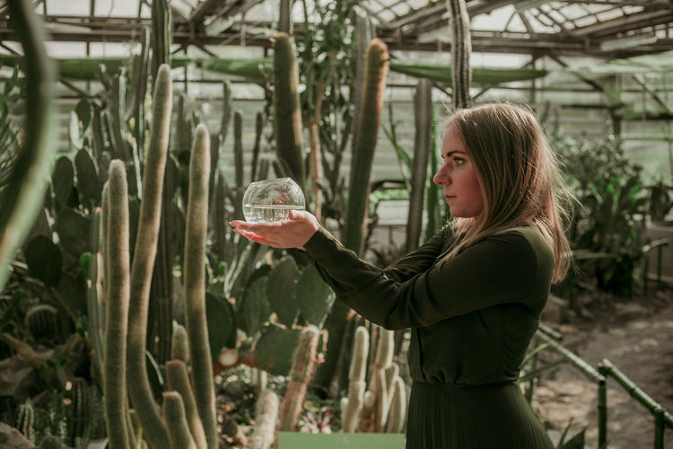 Side view of thoughtful woman holding container while standing in greenhouse