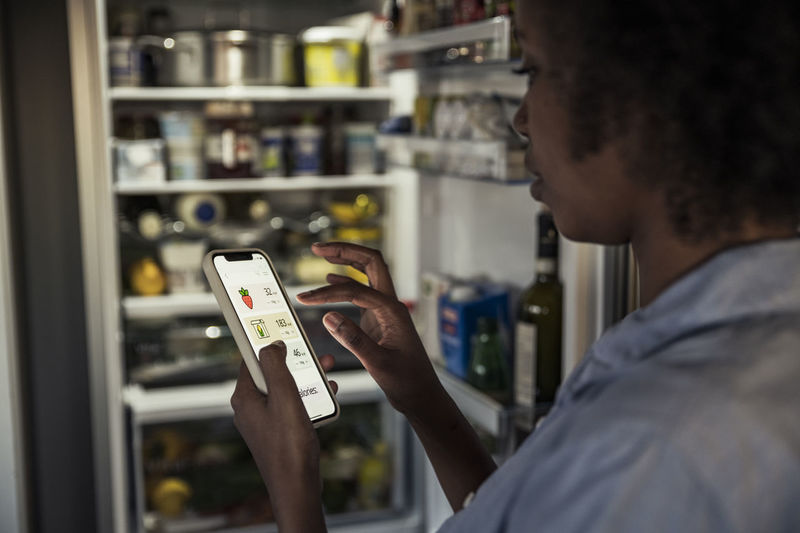 Woman counting food calories through smart phone by refrigerator at home