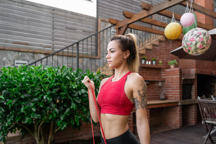 Cheerful female athlete in sportswear doing bicep curl exercise with elastic band during workout in courtyard and looking away