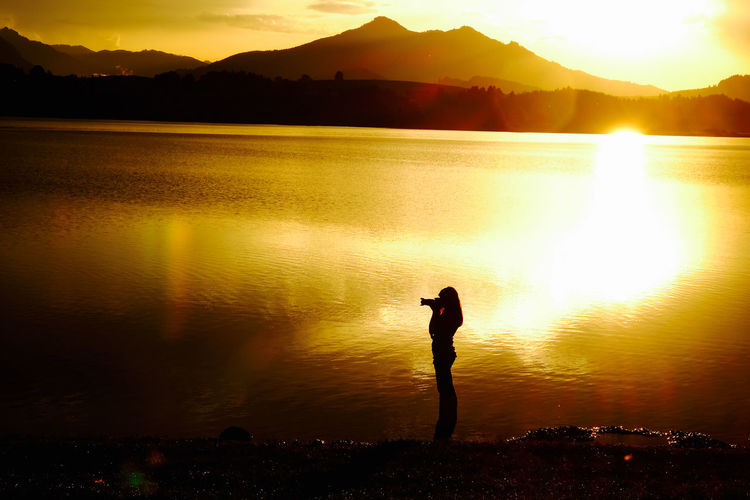Silhouette woman photographing at lakeshore during sunset