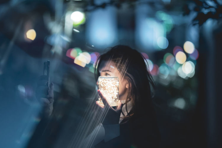 Woman using phone while holding glowing jar in city at night