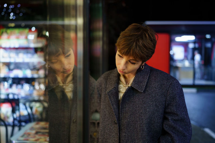 Emotionless caucasian woman with short hair examines shop window standing on street in evening