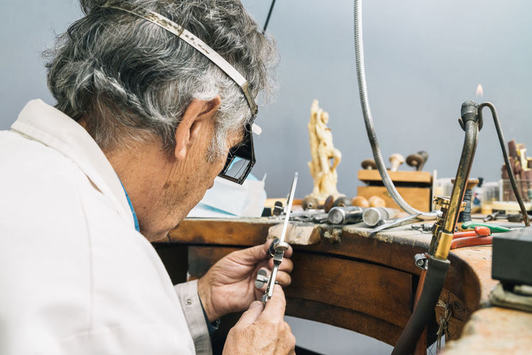 Side view of concentrated skilled mature male master in glasses using ring sizer while creating jewelry in workshop