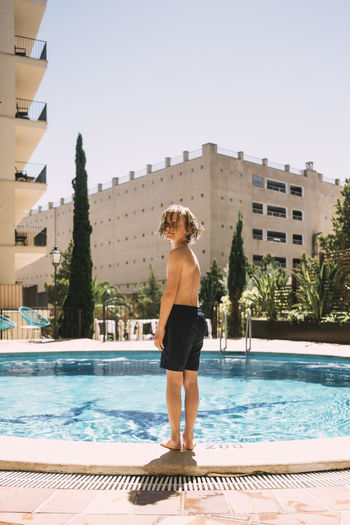 Full length of shirtless boy standing by swimming pool against sky in resort