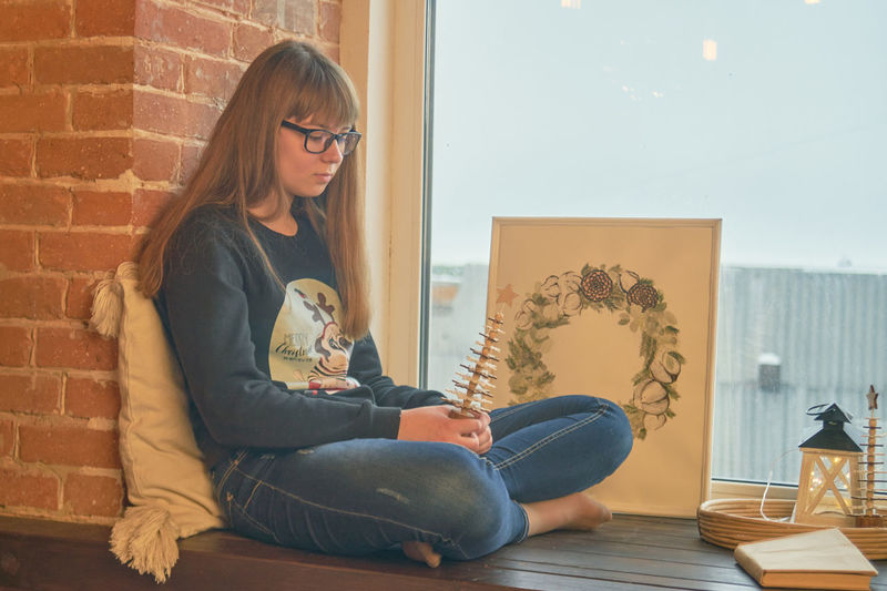 Girl holding decoration while sitting by window against wall
