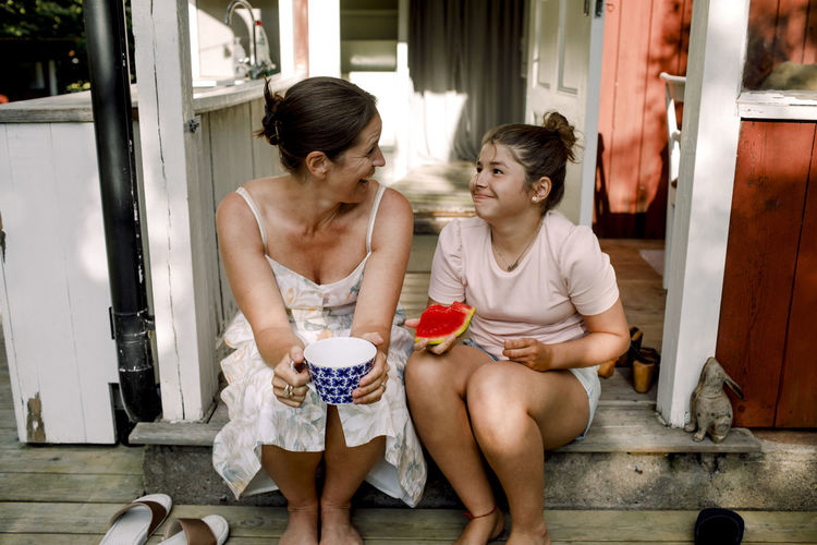 Cheerful mother talking to daughter eating slice of watermelon outdoors