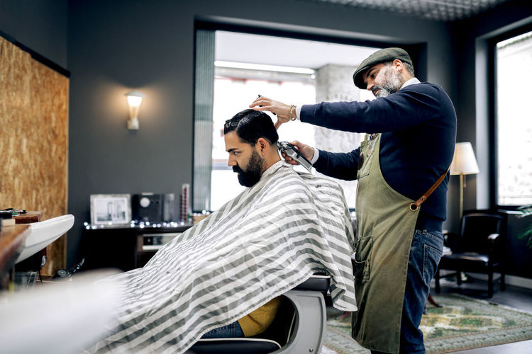 Side view of male ethnic hispanic customer in cape sitting on chair and getting haircut from professional barber working in grooming salon
