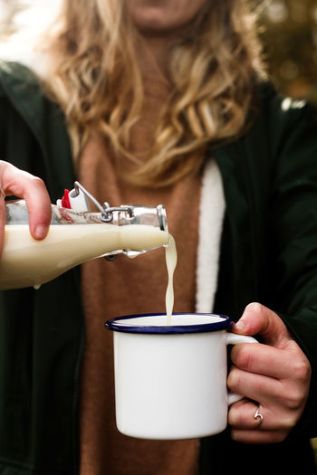 Crop female in warm casual clothes filling white metal cup with tasty dairy beverage from glass bottle while having picnic in nature
