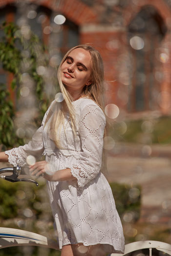Pretty blond girl in white dress with bike near fountain in sunny light