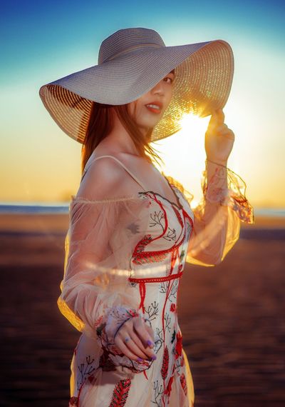 A asian woman wearing a hat standing on the sand with a beautiful sunset background