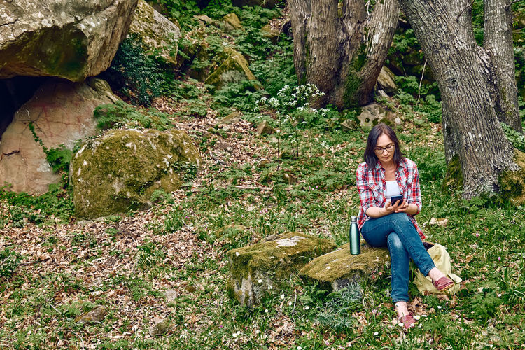 Woman sitting in forest on a stone, using mobile phone for chat or looking for gps.