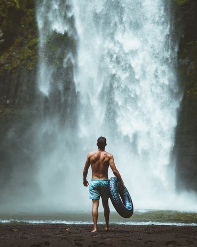 Rear view of shirtless man standing against waterfall