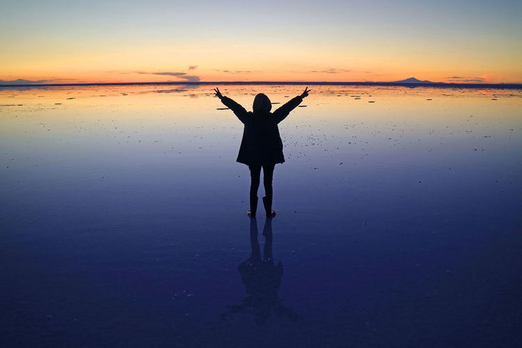 Silhouette of a happy traveler rising arms on mirror effect of uyuni salt flats at sunset, bolivia