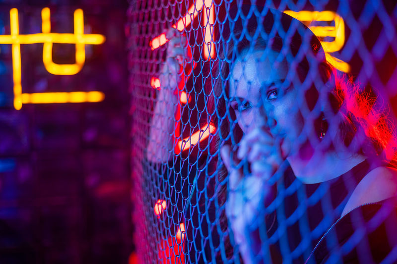 Caucasian woman in neon studio behind chain-link mesh. 