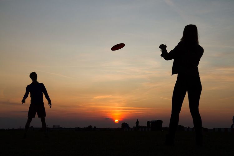 Silhouette man and woman playing with plastic disc on field during sunset