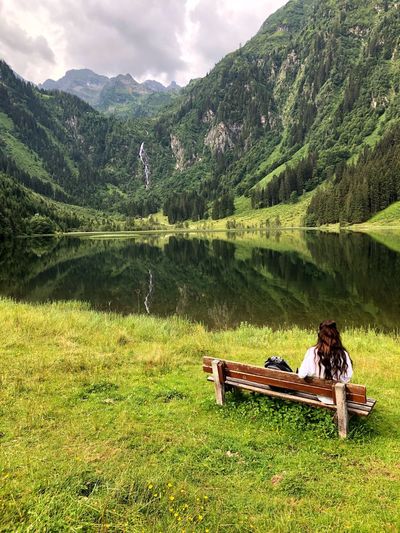 Rear view of woman sitting on bench by lake against mountains