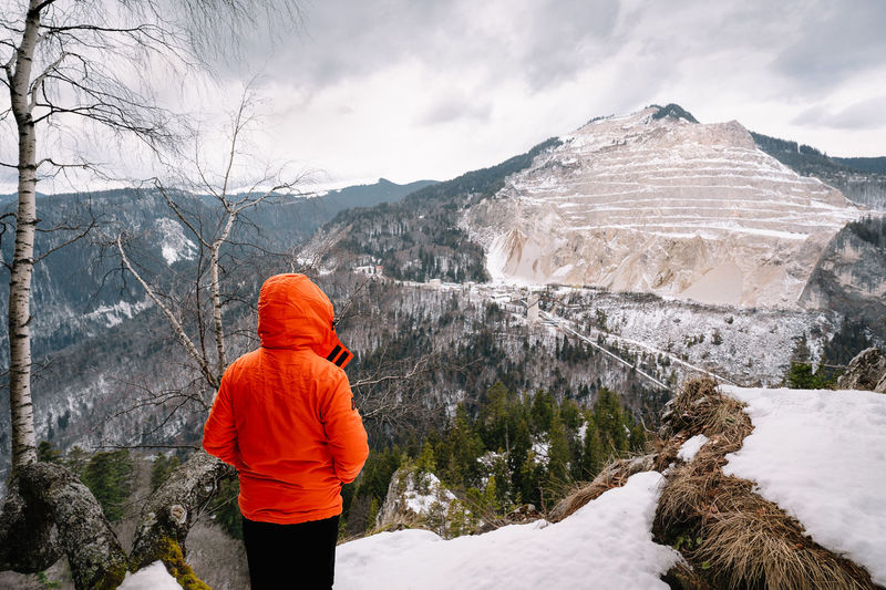 Rear view of person in orange jacket looking at snowcapped mountain during winter