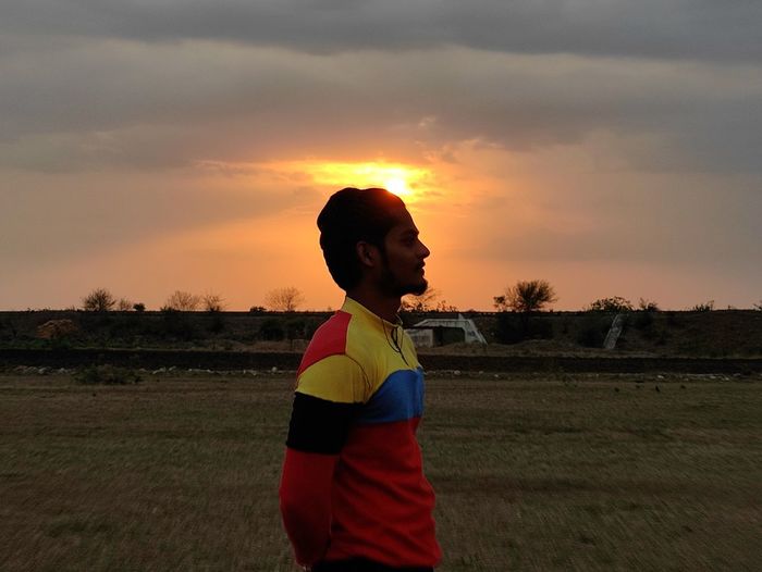 Side view of boy standing on field against sky during sunset