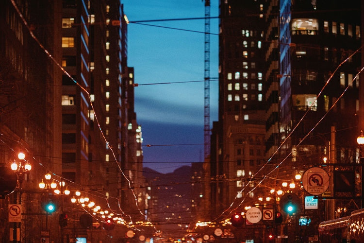 Illuminated street amidst buildings against sky at dusk
