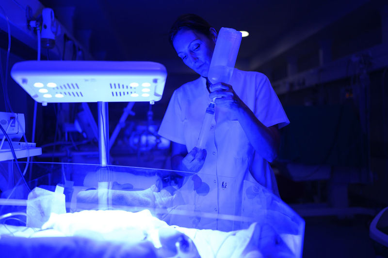 Woman in medical uniform using syringe to take drug from bottle while standing near incubator in neonatal ward with blue illumination during night shift
