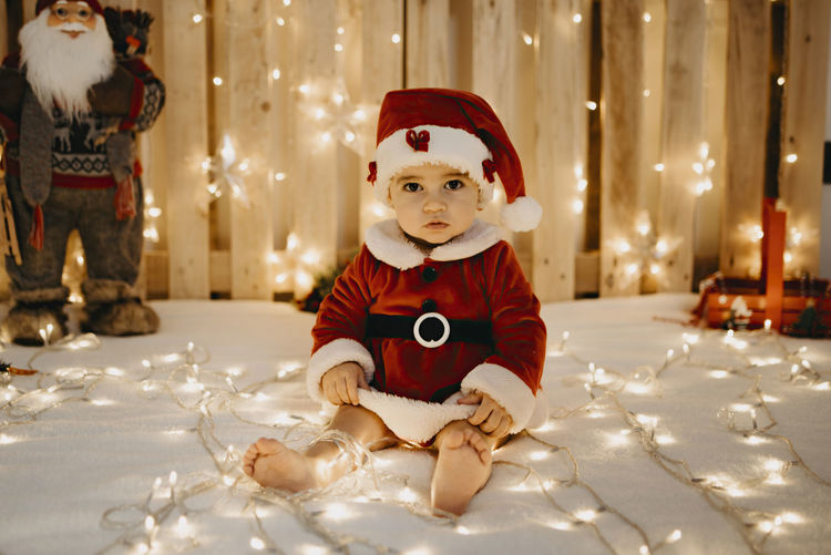 A baby girl sits looking at the camera dressed as a santa claus.