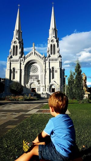 Boy sitting on field by basilica of sainte-anne-de-beaupre against sky
