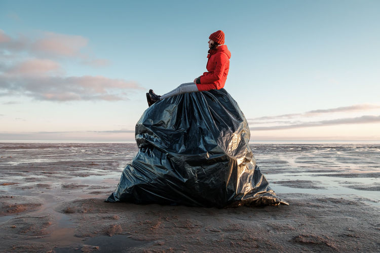 A woman sits on a huge black garbage bag on the shore