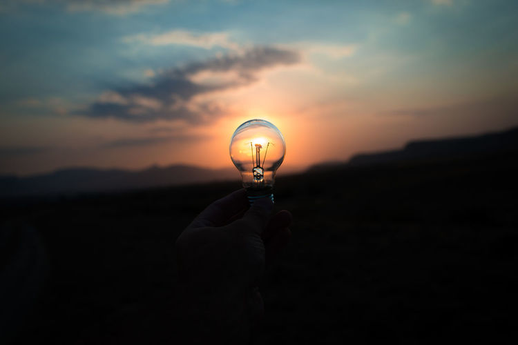 Silhouette hand holding light bulb against sky during sunset