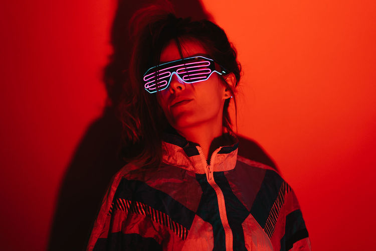 Young female in stylish sunglasses and old fashioned attire standing in studio with red light
