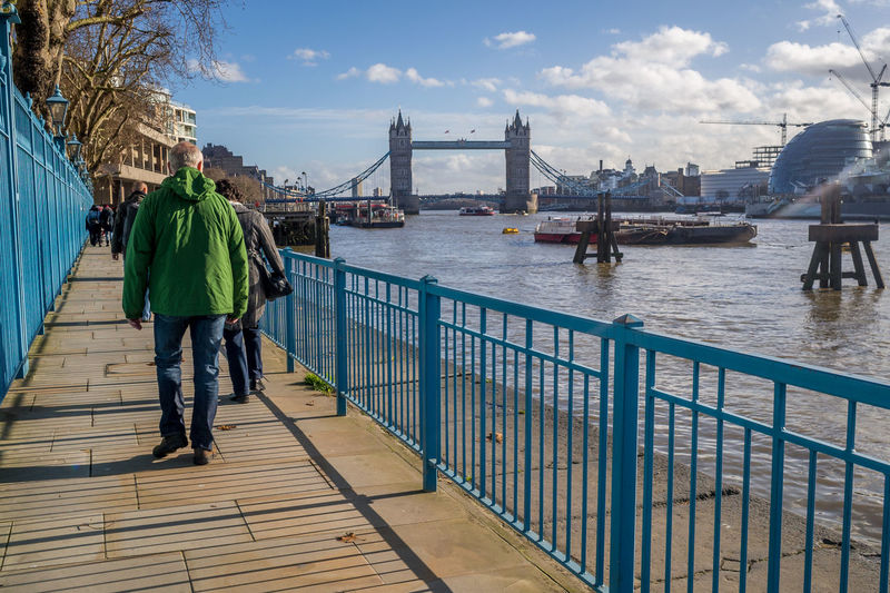 Rear view of people walking on footpath by tower bridge over thames river against sky