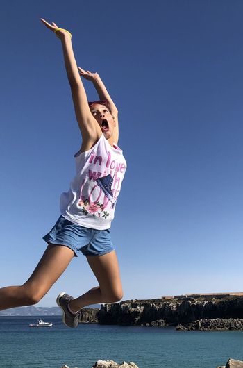 Low angle view of girl with arms raised jumping at beach against clear blue sky