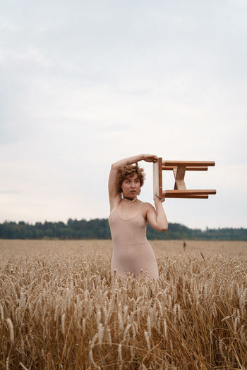 Woman holding stool standing on field against sky