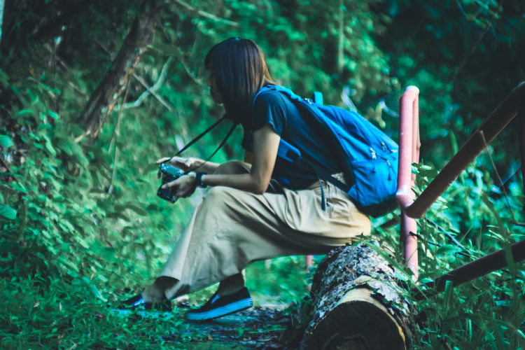 Full length of woman holding camera sitting at forest