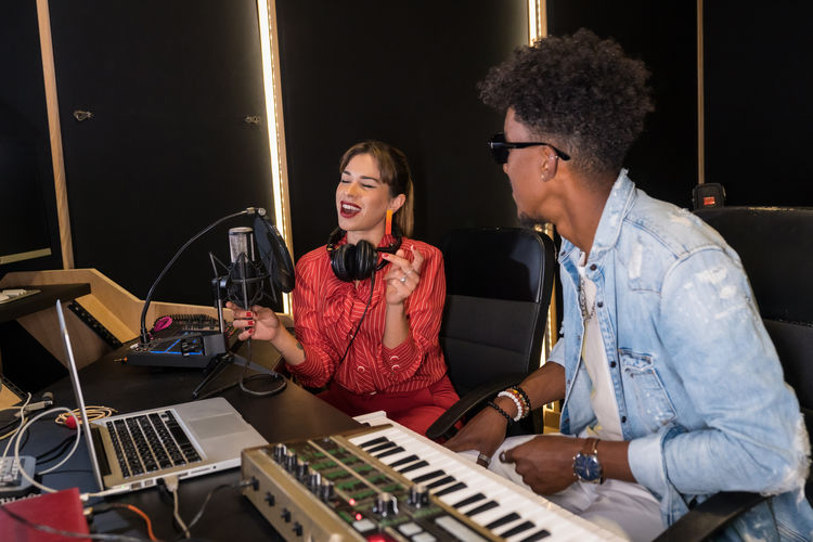 Talented woman singing in mic while recording music in studio with black male musician sitting at table with electric piano