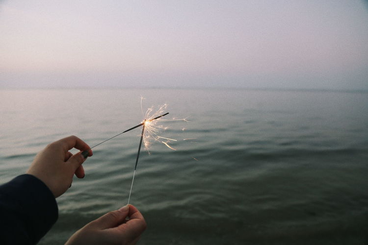 Cropped hands of woman holding lit sparklers over sea against sky during sunset