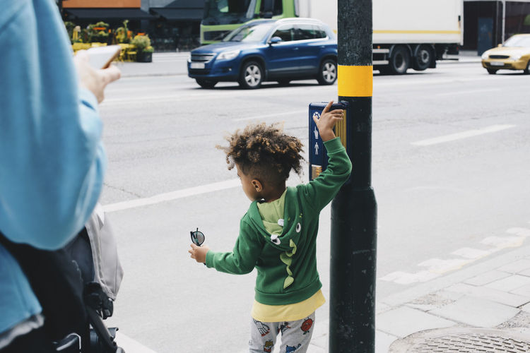 Rear view of boy with mother pushing button on pedestrian crossing sign in city