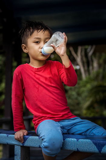 Portrait of boy drinking milk through baby bottle while sitting on railing