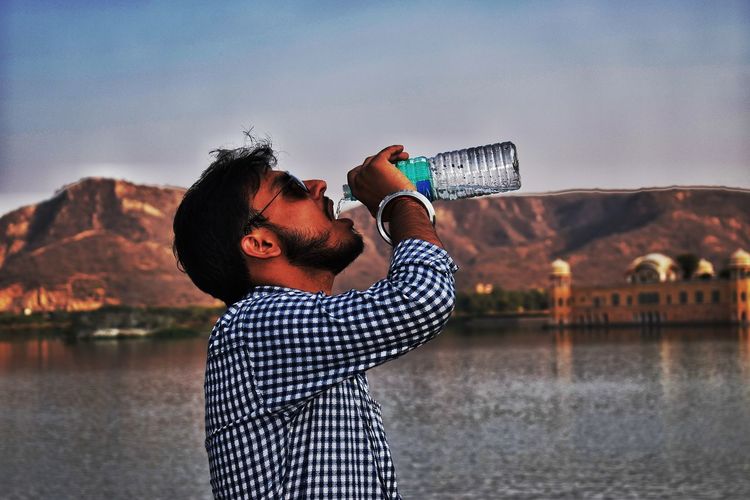 Man drinking water from bottle by lake against sky