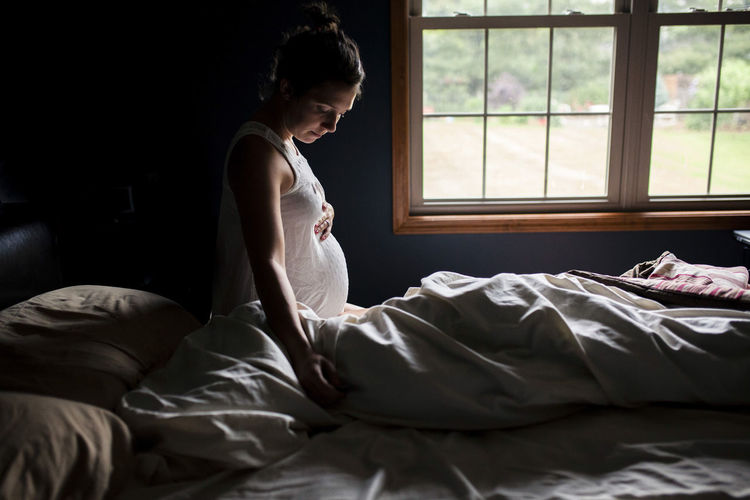 Side view of pregnant woman touching belly on bed at home