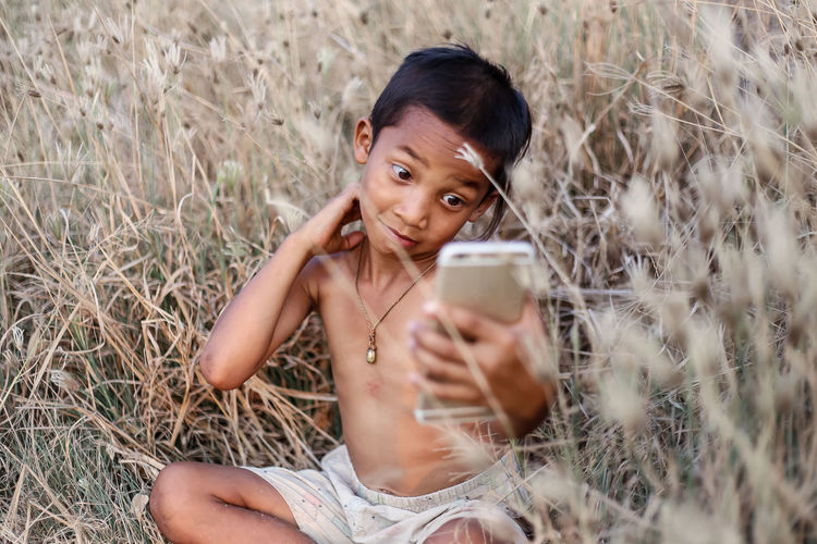 Boy using mobile phone while sitting on field at farm