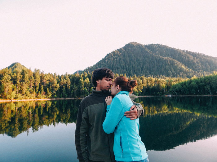 Couple embracing while standing by lake against trees