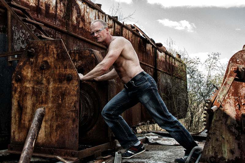 Side view of shirtless mature man exercising on abandoned metal against sky