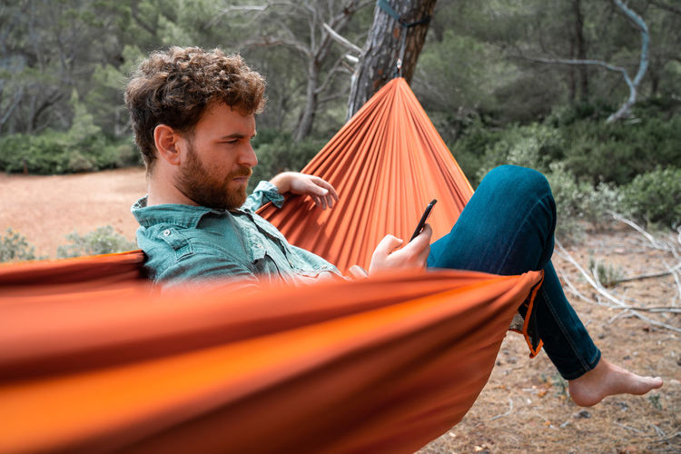 Full length of man sitting using smart phone while on hammock in forest