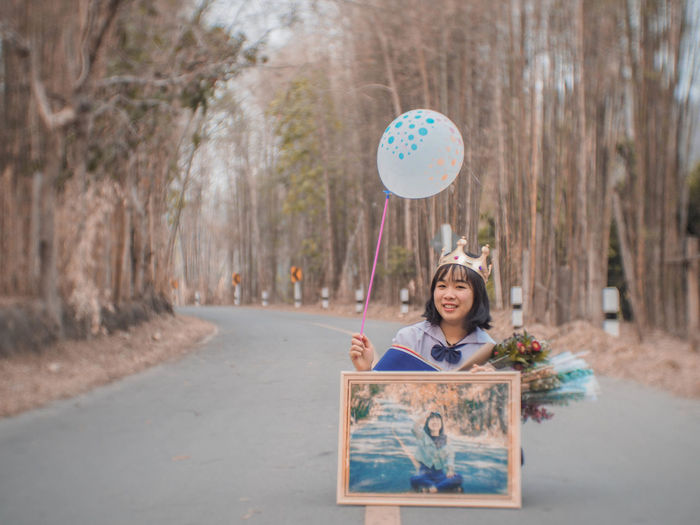 Portrait of young woman holding balloon while standing on road