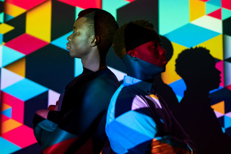 Side view of emotionless african american men standing back to back in studio with colorful neon illumination in shape of cubes