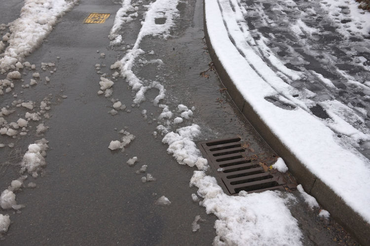 Surface drain on the corner of a tarmac road, surrounded by melting snowfall. winter environment