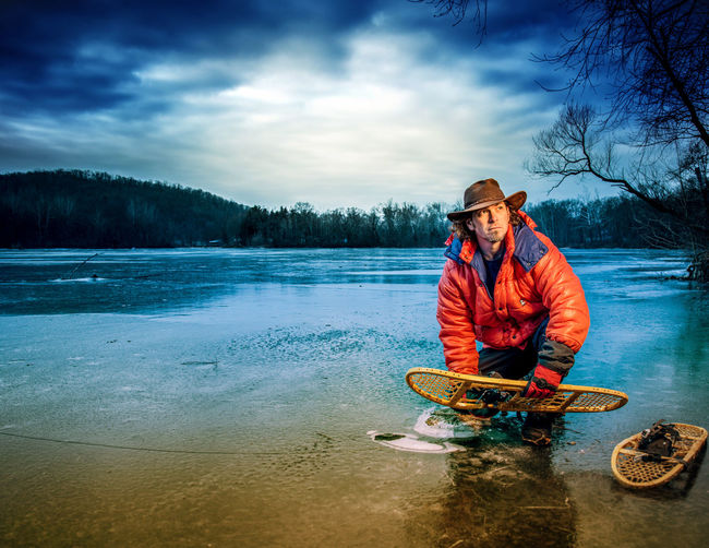 Full length of man holding snowshoes in river against cloudy sky at dusk